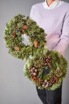 Christmas wreath of fresh spruce in woman hands. Xmas circlet with ornaments, toys and balls. Christmas mood. Gray wall on background
