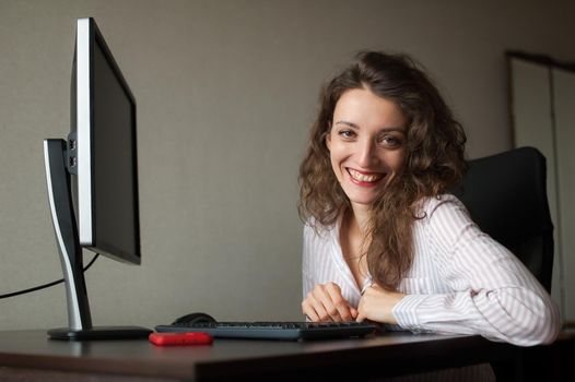 Young female office manager in white shirt and curly hair is sitting at the table and typing using keyboard, routine work, freelance.