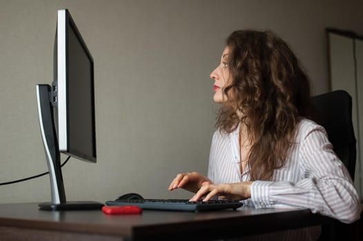 Young female office manager in white shirt and curly hair is sitting at the table and typing using keyboard, routine work, freelance.