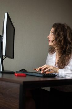 Young female office manager in white shirt and curly hair is sitting at the table and typing using keyboard, routine work, freelance.