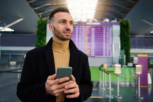 Handsome Caucasian young man using smartphone and checking flight standing in front of check-in board at departure terminal of international airport