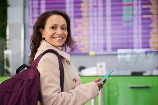 Close-up of beautiful Middle Eastern woman with backpack using mobile phone, browsing websites, making hotel reservations, smiling toothy smile standing at flight information panel in airport terminal