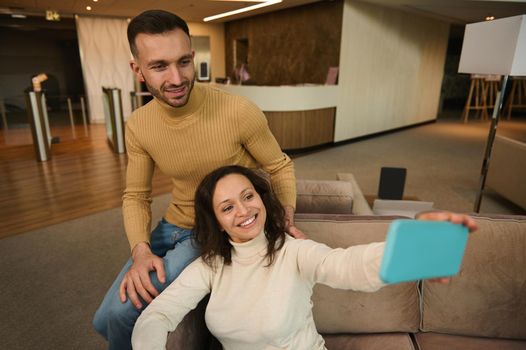 Attractive Hispanic woman sitting next to her boyfriend and making a self-portrait on smartphone while resting together in the lounge zone of the airport departure terminal while waiting for a flight