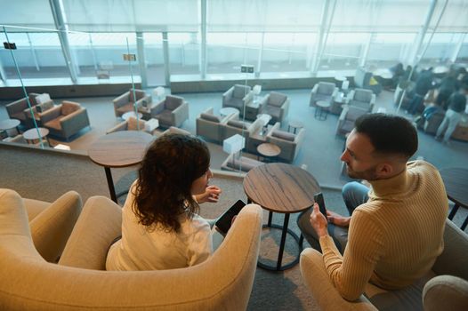 Overhead view of a young contemporary multiracial couple in love, at honeymoon travel discussing forthcoming flight while waiting for airplane departure, sitting at table in the airport lounge