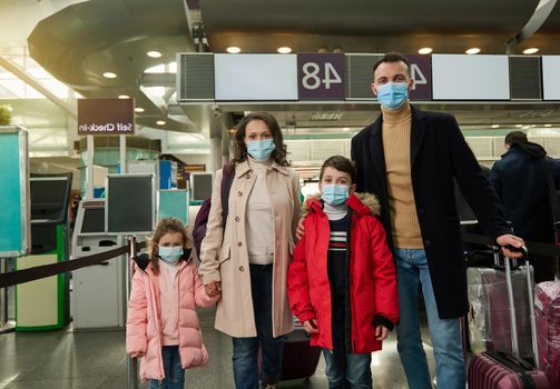 Portrait of a young family wearing protective medical masks traveling during a pandemic, standing with luggage and suitcase at the airport while waiting to check in for a flight. Air travel concept