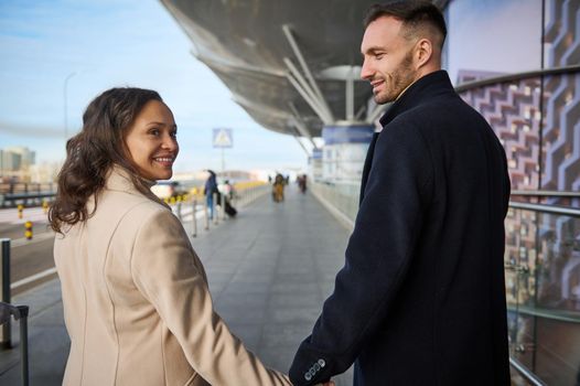 Loving young reunited multiracial couple looking at each other, smiling, holding hands while walking along the road of the international airport in the arrivals area