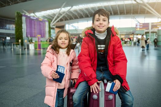 Happy cheerful European kids- boy and girl in warm clothes, sitting on a suitcase and holding passports, with air tickets and boarding pass at the international airport. Family airplane travel concept