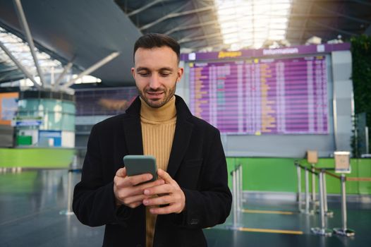 Handsome smiling Caucasian young man, business traveler using smartphone, booking hotel, standing with his back to flight information panel with timetable in arrivals terminal of international airport