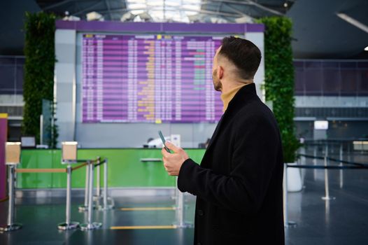 Handsome young Caucasian man, businessman on a business trip, dressed in a dark coat, with a phone in his hands, looks at the information panel of the timetable at the international airport