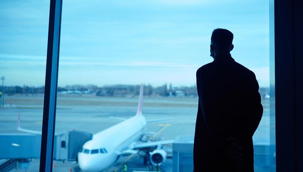 Rear view of a silhouette of a businessman during business trip standing by the panoramic window in the airport lounge, looking at the plane and the runways while waiting for his flight
