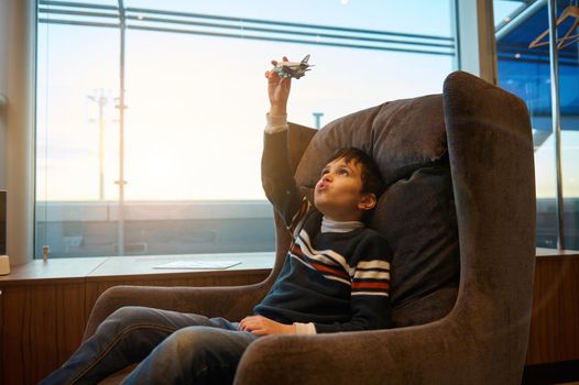Handsome European teenage boy plays with toy airplane sitting in a chair by panoramic windows overlooking the runway at sunset while waiting to board flight at international airport departure terminal