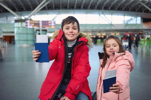 Handsome teenage boy and cute little girl wearing warm down jackets smiling toothy smile while standing at international airport and holding passport documents with air tickets and boarding pass