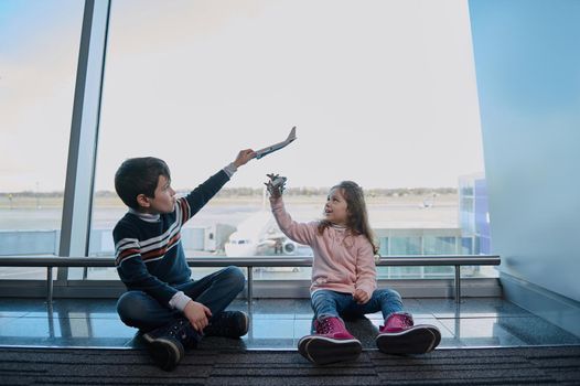 Handsome boy and girl, brother and sister playing with toy airplanes sitting on the floor by the panoramic windows of the airport departure terminal overlooking the runways. Family air travel concept
