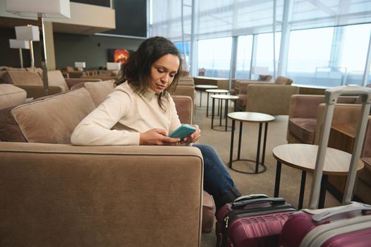 Confident business woman traveling alone, sitting at a table and using a mobile phone, relaxing on an armchair in the luxurious lounge of the airport departure terminal while waiting to board a flight