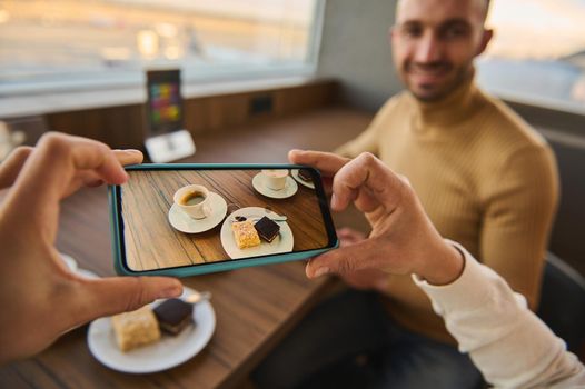 Woman hands taking photo on smartphone of cup of coffee and cakes on wooden table with blurred handsome man sitting at panoramic windows overlooking the planes on runways. Mobile phone live view mode