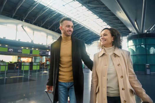 Couple in love with luggage waiting for check-in, walking through the departure hall of airport terminal. Loving Caucasian man and woman flying on vacation for honeymoon. Safe travel after a pandemic