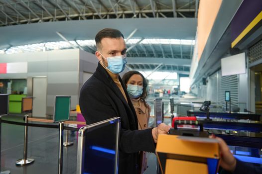 Young couple of business partners in protective medical masks, traveling during pandemic, standing at the check-in counter, passing customs and passport control at the airport terminal. Copy ad space