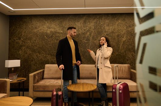 Delighted newlywed couple in the VIP lounge of the airport terminal. Handsome business man looks at his partner who is on the phone, negotiating and booking a hotel while waiting to board a flight