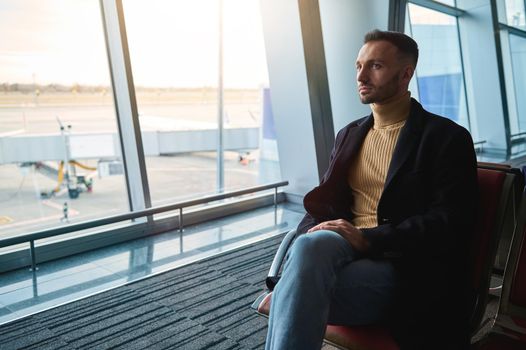 Handsome attractive man, confident businessman on business trip, sitting in the departure lounge, admiring the view from the panoramic windows of the international airport to the runway with airplanes