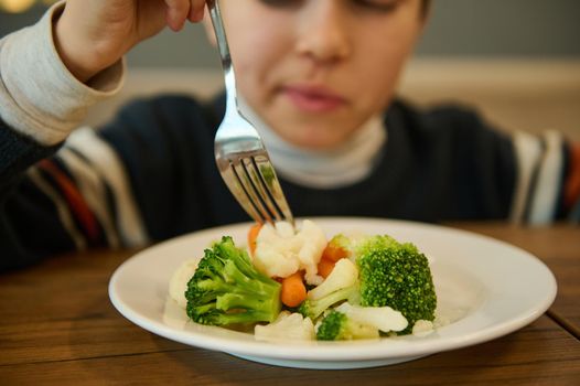 Nutrition, vegetarian diet for toddler. Focus on a plate of steamed vegetables, healthy meal of broccoli and carrots against blurred boy eating sitting at a table and holding fork