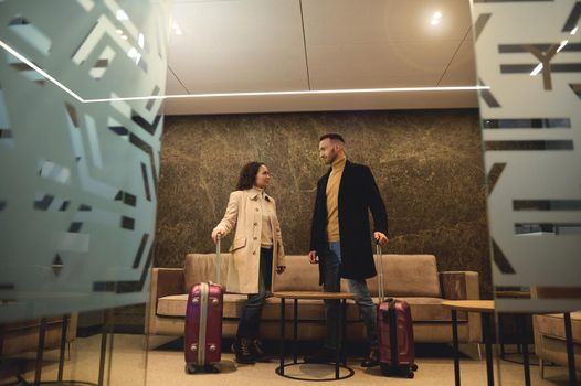 Handsome attractive businessman talking to his business partner, beautiful woman standing with suitcases at the exit from the conference room in the airport terminal ready to board the flight together
