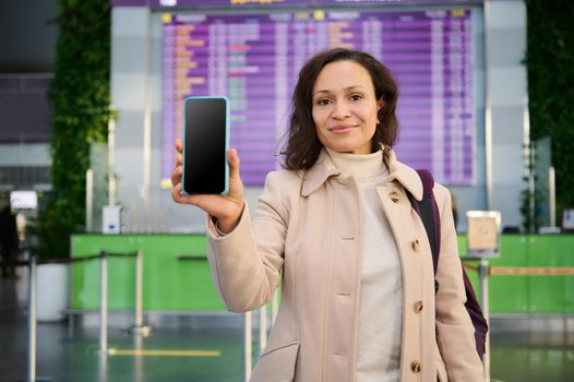 Beautiful woman showing her mobile phone with blank screen to camera, standing by flight information panel and check-in board in departure hall of airport terminal. Copy space for mobile applications