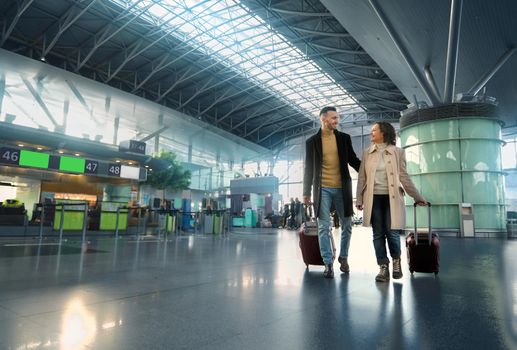 Full-length portrait of a loving couple travelling together, man and woman with suitcases walking along the departure hall of international airport on a blurred background of flight check-in counters