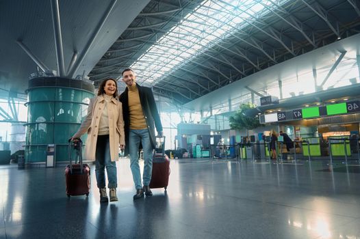 Beautiful couple in love enjoys a joint journey, walks through the departure-arrival hall, waits for customs and passport control at an international airport on the background of flight check-in board