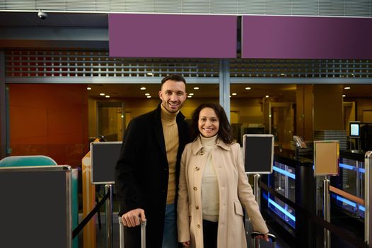 Portrait of two people, multiracial couple, beautiful Arab woman and Caucasian handsome man with suitcases smiling toothy smile while going through customs and passport control at the airport terminal
