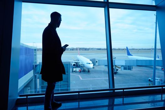 Silhouette of businessman in airport lounge with mobile phone standing by panoramic windows overlooking runways and airplanes waiting for flight early in the morning. Business trip, air travel concept