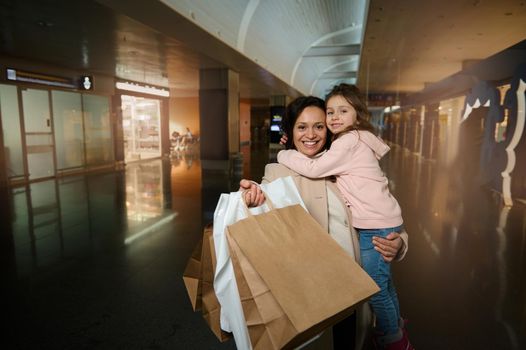 Happy mother holding and hugging her cute daughter in arms, smiling, looking at camera showing shopping bags standing in duty free area of departure international airport terminal, waiting for flight