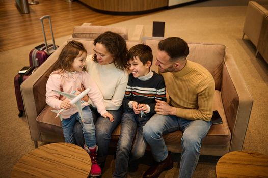 Happy family. Parents admiring their kids playing with toy planes, discussing the upcoming air trip, resting in the VIP lounge of the airport departure terminal while waiting to board the flight