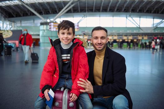 Caucasian man- father and adorable preadolescent son in red down jacket sitting on suitcase holding passport and boarding pass smiling looking at camera in departure area of international airport