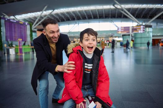 Cheerful happy young father rides his son on a suitcase in the departure hall of the international airport. Dad and son having fun together while waiting for passport and customs control and boarding