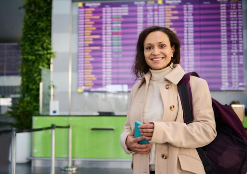 Attractive Middle Eastern woman with backpack and mobile phone, smiling with beautiful toothy smile looking at camera standing at flight information panel and flight check-in board in airport terminal