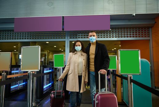 Young couple of business partners in protective medical masks, standing with suitcases at check-in counter, waiting for check-in, passing customs and passport control in airport terminal. Copy space