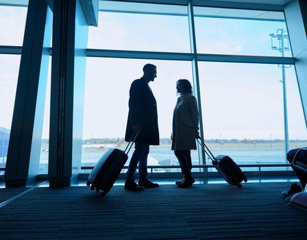 Silhouette of two people, woman and man with suitcases, standing by the panoramic windows overlooking the runways and planes in the departure terminal of the international airport, awaiting a flight