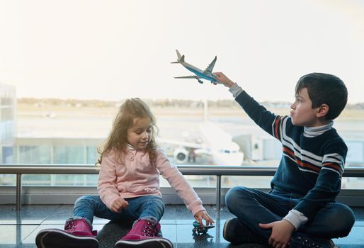 Handsome kids, boy and little girl playing with toys planes sitting on the floor by the panoramic windows overlooking the runways and planes in the departure terminal of the international airport