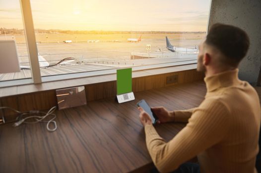 Rear view. Businessman entrepreneur handsome man with smartphone sitting at table by panoramic windows with view of airplanes on runway at sunset, rests in airport lounge cafe while waiting for flight