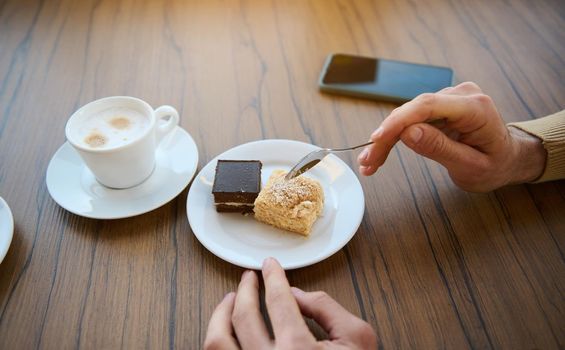 Close-up. Crop. Man's hands holding tea spoon near delicious sweet dessert, chocolate and cheese creamy cake, cup of aroma coffee with foam and smartphone on wooden table. Coffee break, snack concept