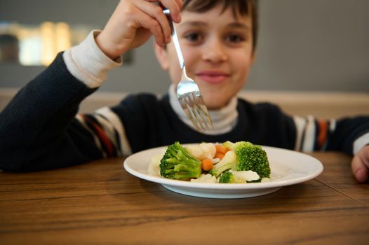 Image focused on a plate of steamed vegetables. Adorable smiling Caucasian teenage boy sitting at table and eating healthy lunch of steamed carrot and broccoli. Nutrition, vegetarian diet for toddler