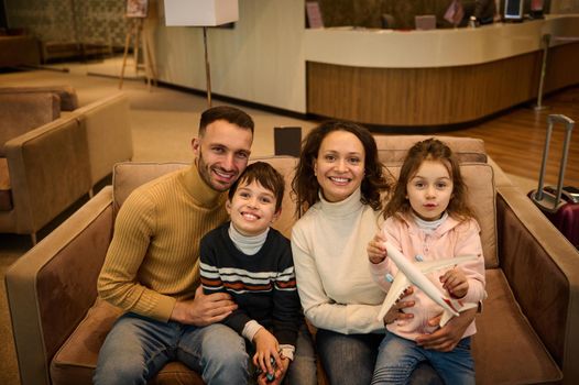 Happy multi ethnic family- mom, dad, son and daughter with toy airplane sitting on sofa in VIP lounge, smiling with cheerful toothy smile looking at camera waiting for flight in the airport terminal