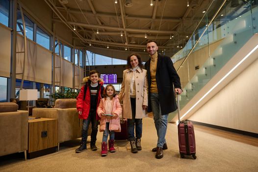 Delighted happy kids and parents enjoying family travel, standing with suitcases in a lounge zone of airport departure terminal against the flight information panel with schedules. Post pandemic trip