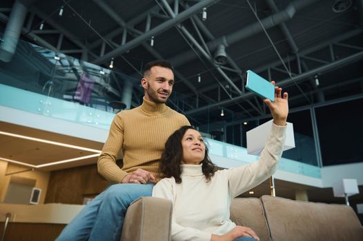 Charming woman and handsome man, friends, newlyweds, multiracial heterosexual couple in love taking selfie while waiting to board a flight in an international airport lounge during honeymoon trip.