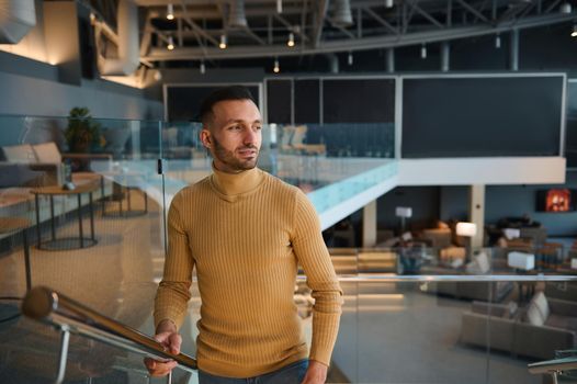 Handsome attractive Caucasian confident man, businessman on business trip climbs the stairs in waiting room of departure terminal at international airport, looking away while waiting for the flight