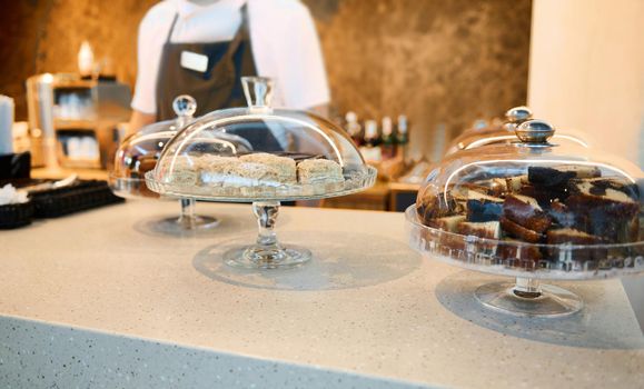 Image focused on cloche with pastries and chocolate cheese creamy desserts and cakes on a restaurant countertop with blurred chef in white uniform and black apron on background. Food and drink concept