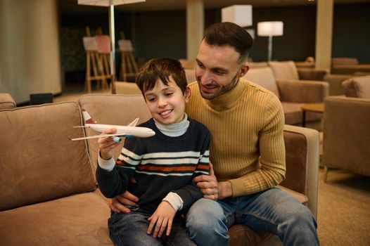 Adorable little boy plays with a toy plane while his loving father sits next to him, hugs him, waiting for boarding a flight in the VIP lounge of the departure terminal of the international airport