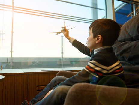 Portrait of adorable child boy holding a toy airplane in outstretched hand imitating flight, against panoramic windows overlooking the runway in the airport departure terminal while waiting for flight