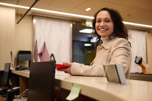 Charming business woman standing with a mobile phone in her hand at the check-in counter in the VIP lounge of the international airport departure terminal, smiling cutely at camera, waiting for flight