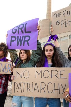 Young woman looking at camera holding No more patriarchy banner in feminist demonstration for woman's rights amongst other protesters. Vertical image. Woman empowerment concept.